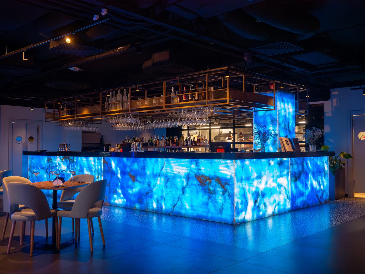 VODA's bar after dark: a blue-lit counter front with a water-like texture and a brass rack of glasses above