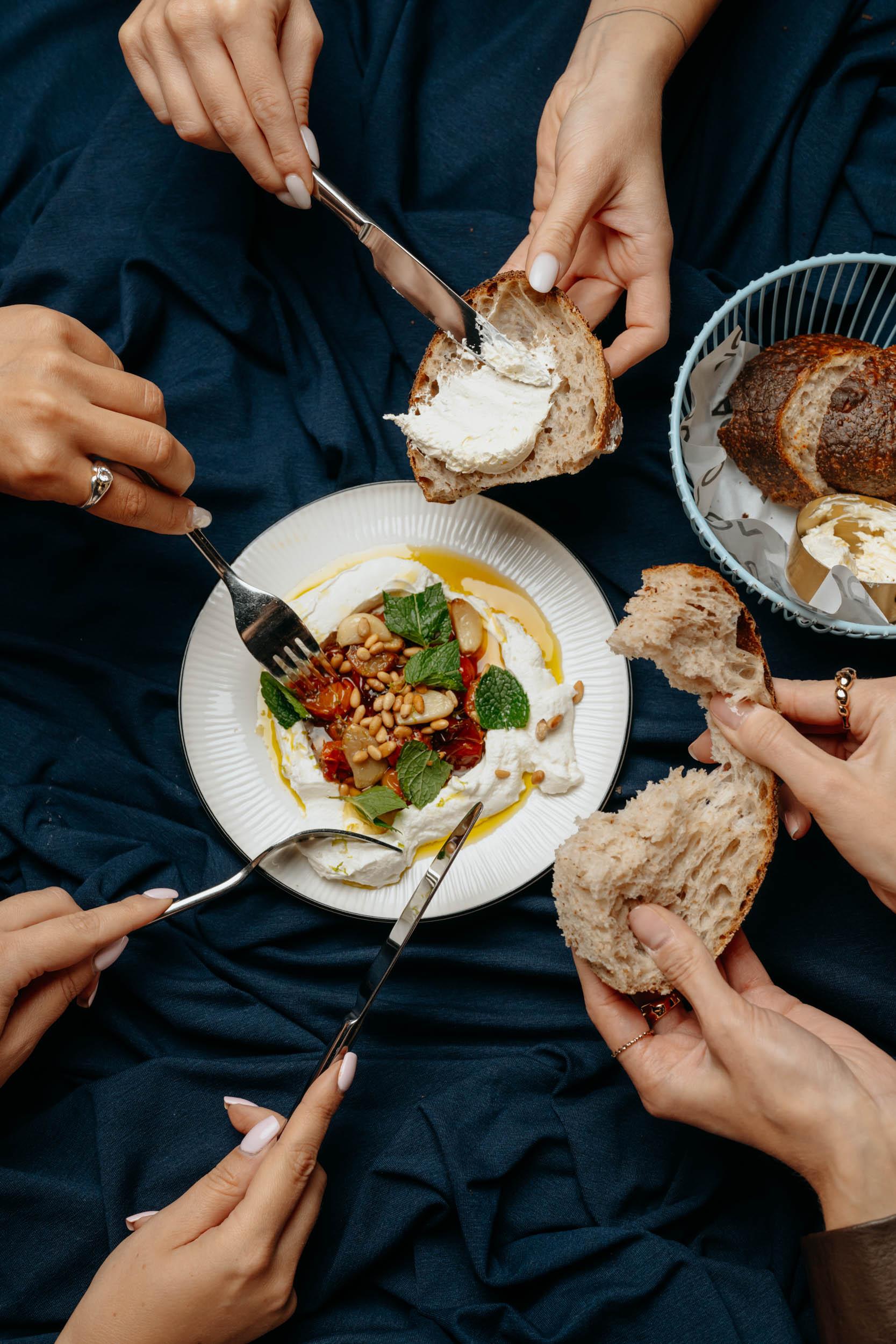 Five pairs of hands over a shared plate of white cheese, roasted tomatoes and mint, with a bread basket beside it