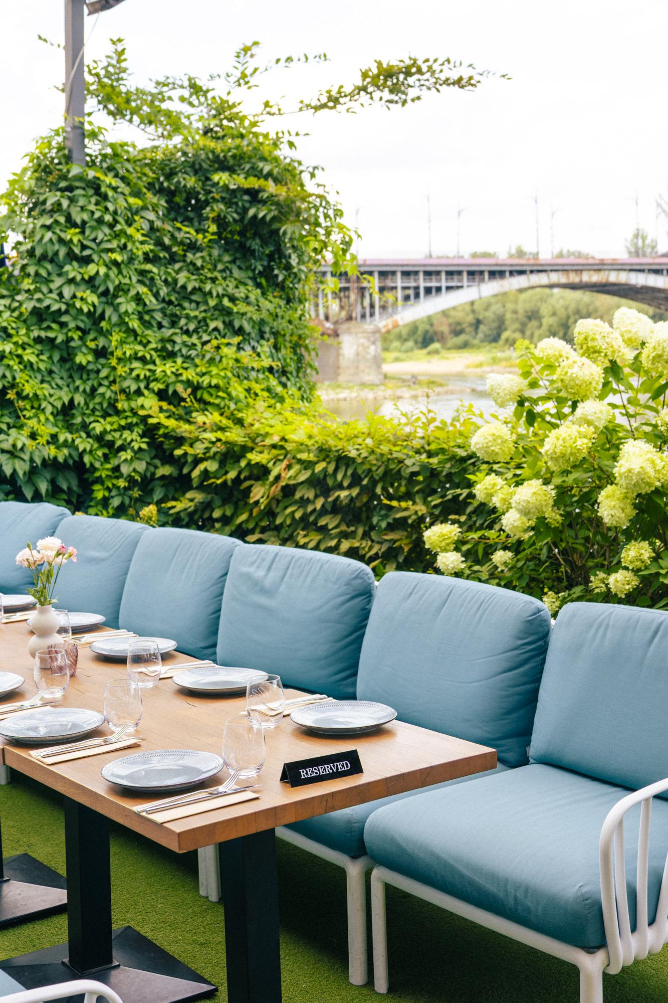 VODA's garden in daylight: pale blue sofas at set tables, flowering hydrangeas and a green hedge, with the Poniatowski Bridge over the Vistula behind