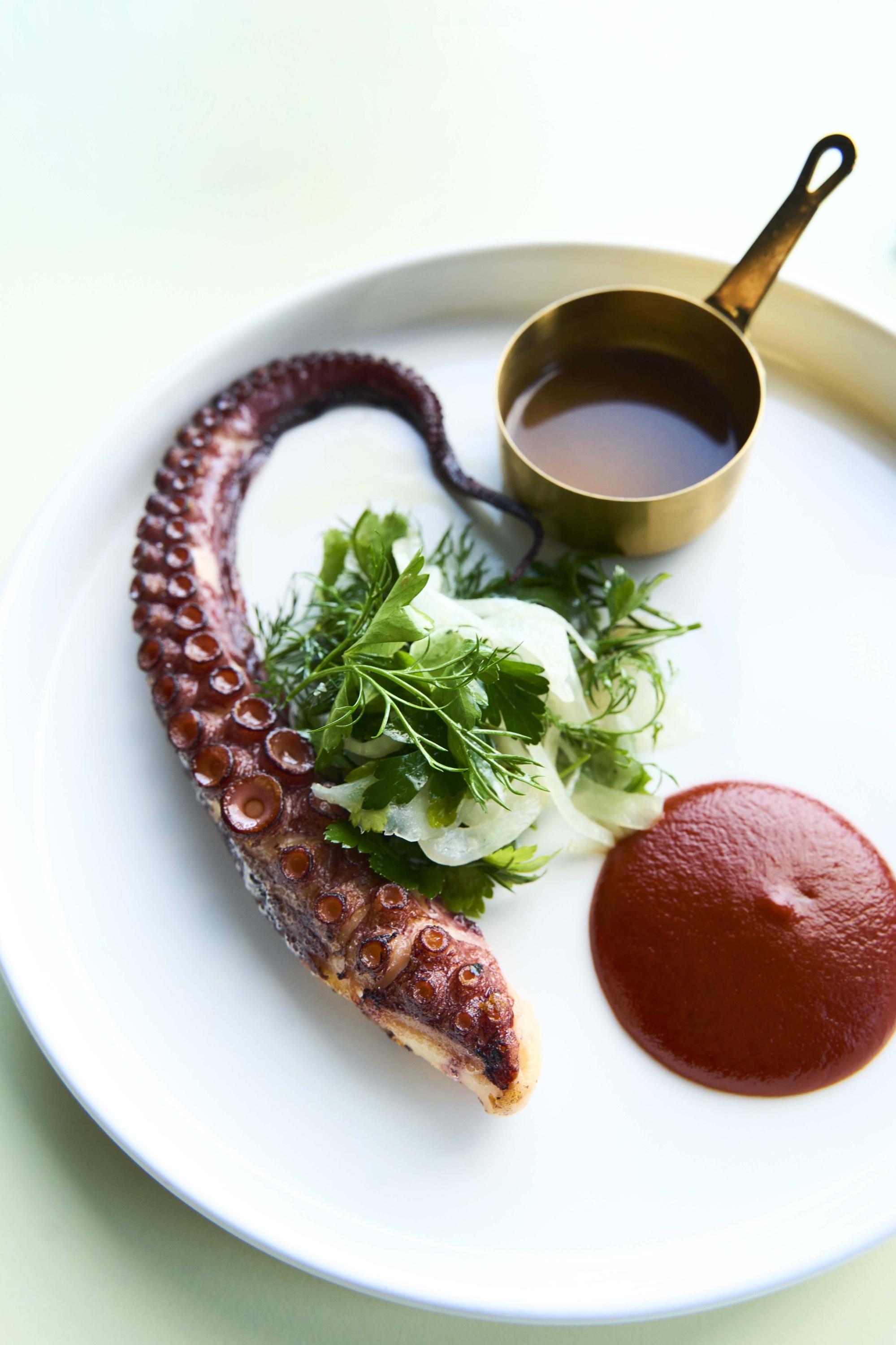 An octopus tentacle on a white plate with herbs, a dark swirl of sauce and a small brass pan of sauce