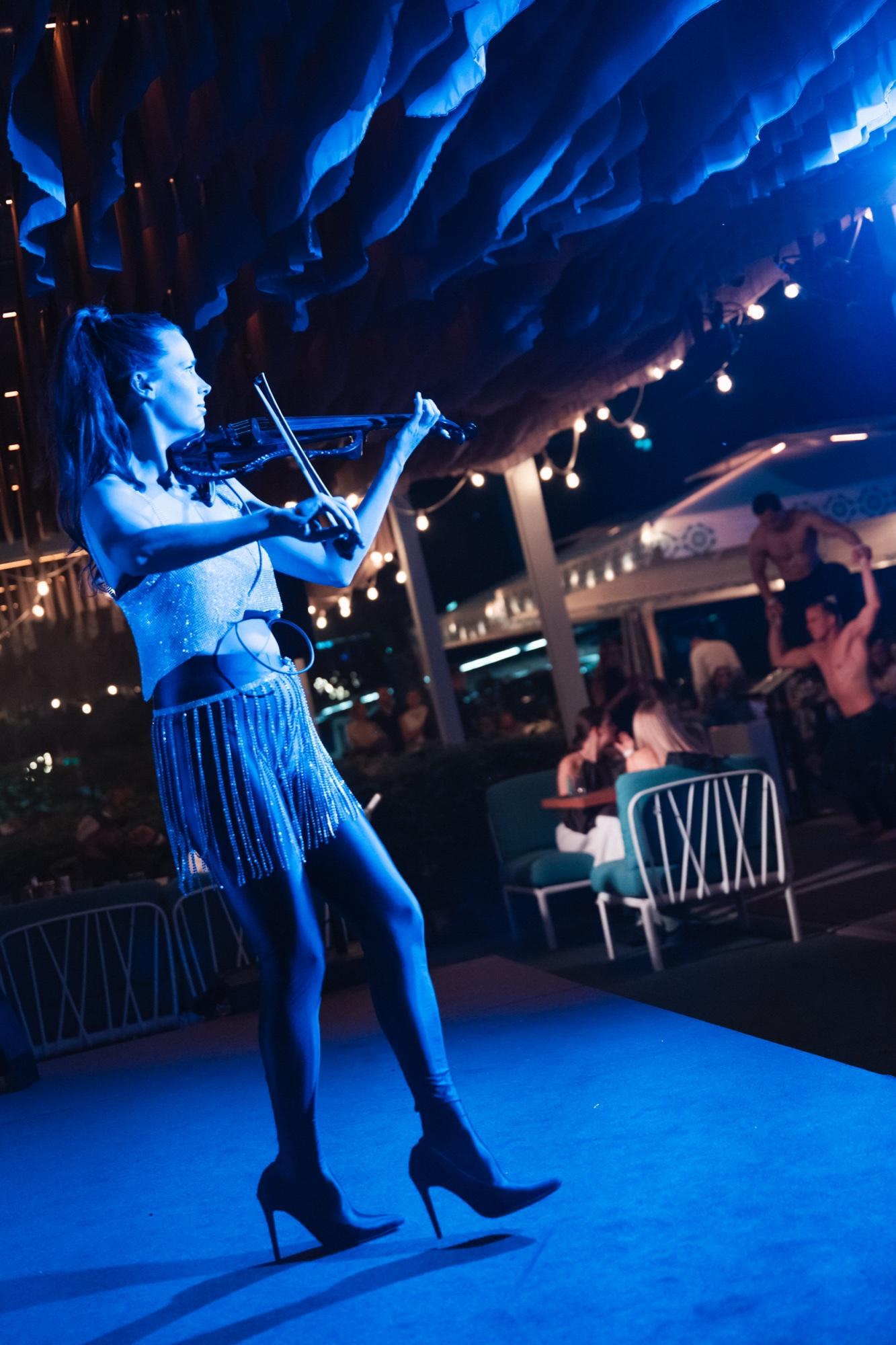 A violinist in a silver costume playing on a blue-lit runway, with guests at tables behind her.