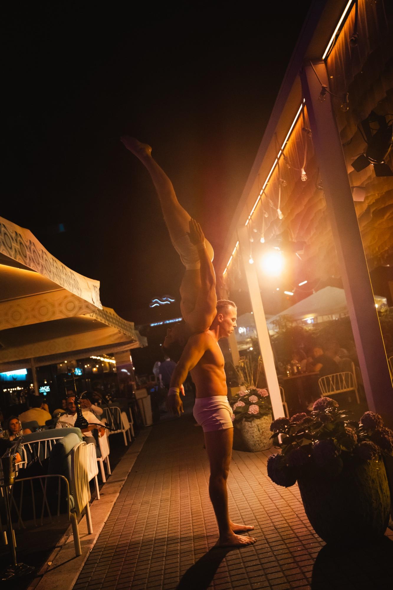 An acrobat in a handstand on the terrace in the evening, next to tables of guests and warm lamplight.