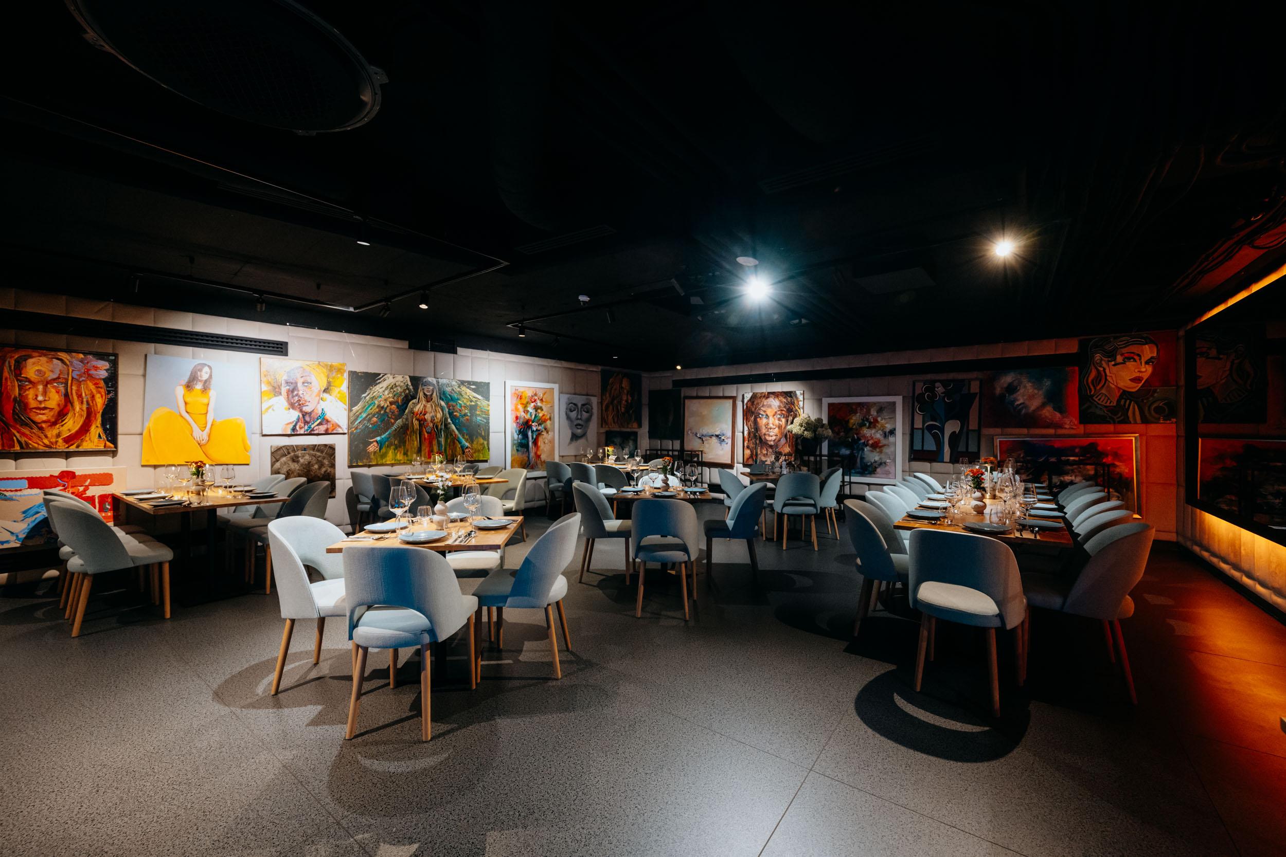 VODA's dining room in the evening from a wide angle: set tables, pale blue chairs and a row of colourful paintings on the wall