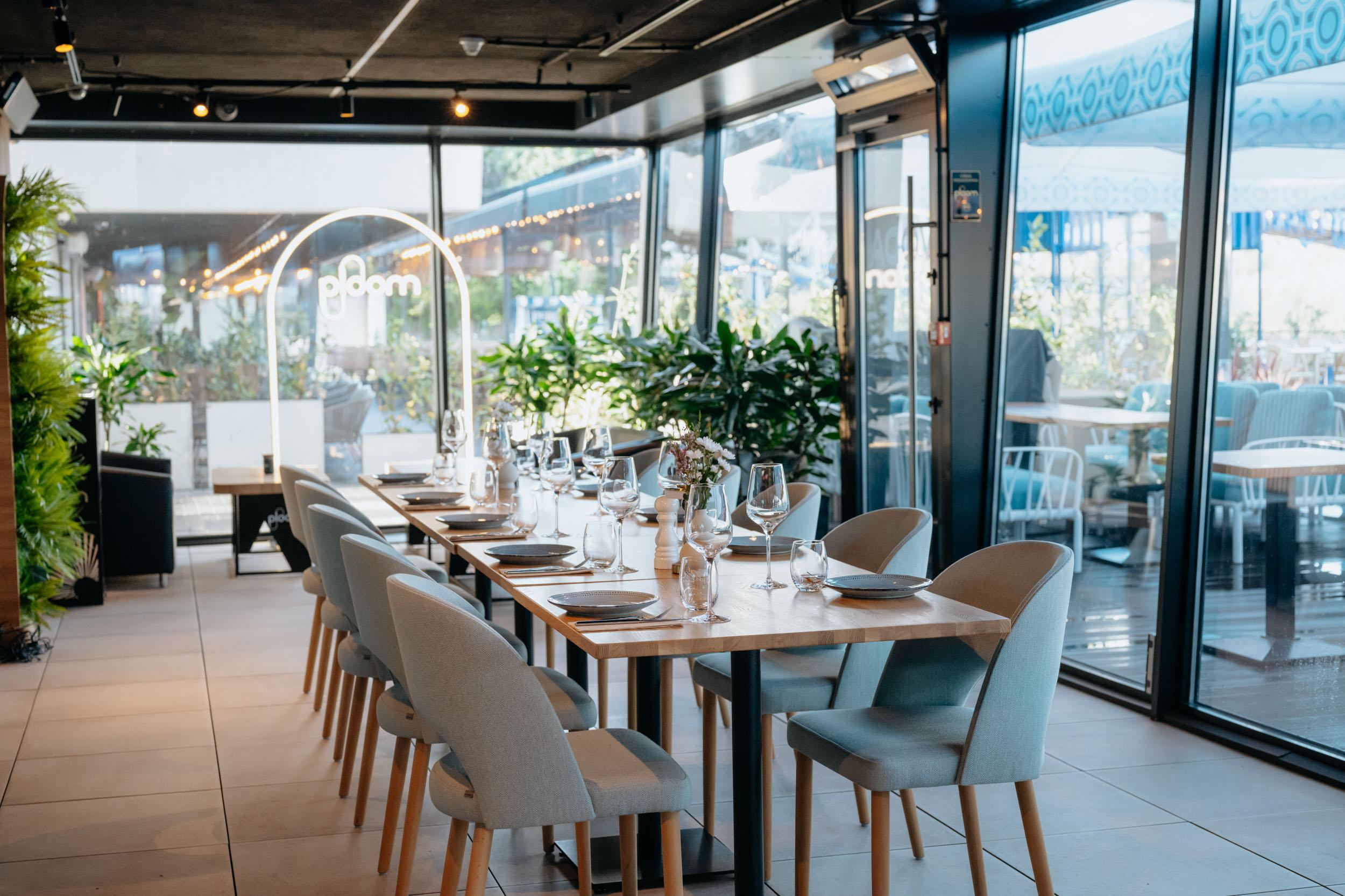 VODA's glazed terrace in daylight: a long table set with glasses and plates, pale blue chairs, plants in planters, the garden beyond the glass